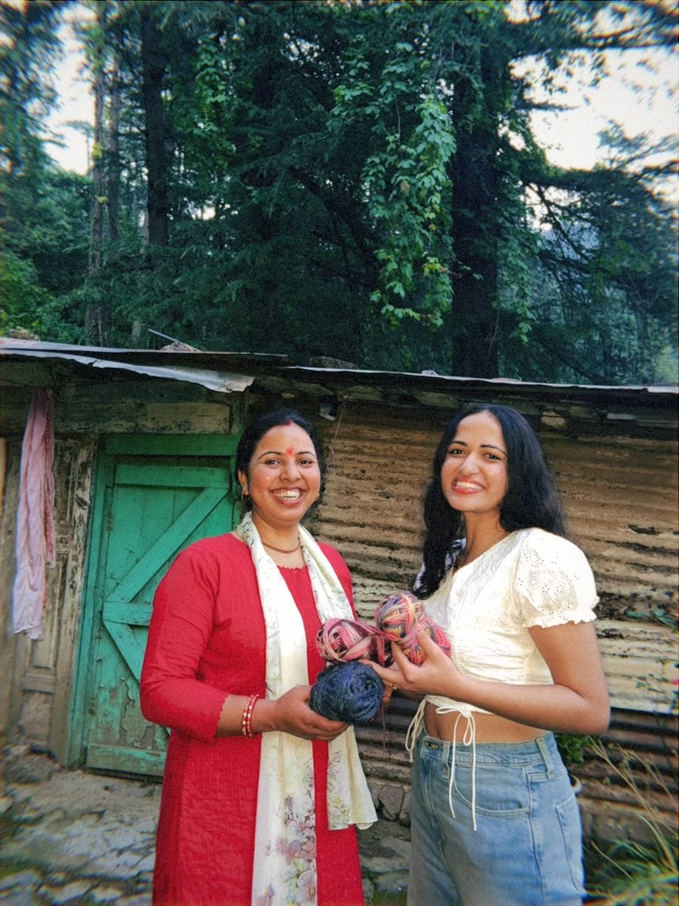 Two women holding yarn in front of a wooden home
